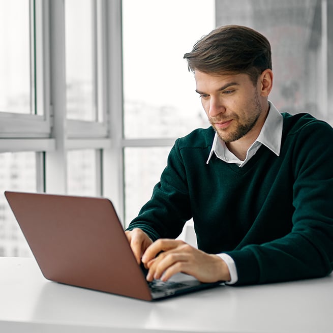 A man sitting by a window in a highrise building typing on a laptop.