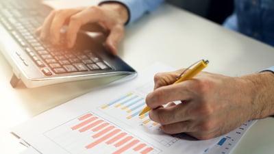 A close up shot of a man typing on a keyboard and taking notes on a piece of paper with data