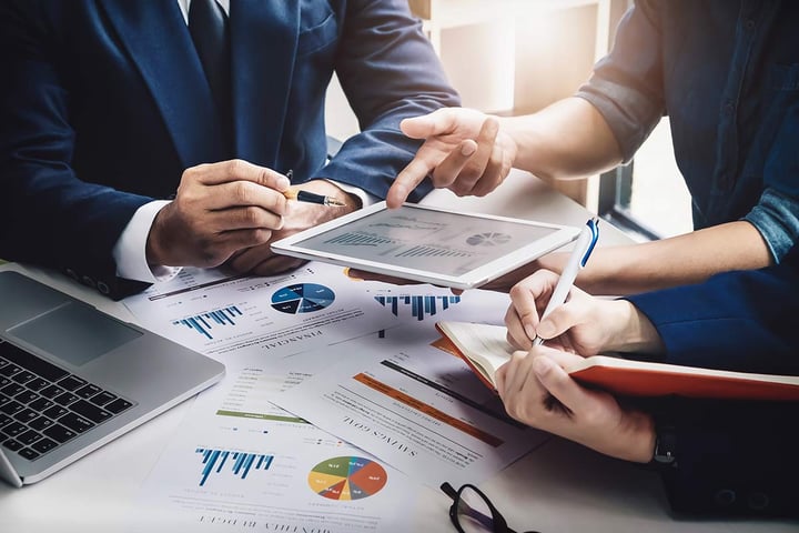 Three business people at a table covered in papers that have graphs and statistics. One person is holding a tablet and pointing at the data on it.